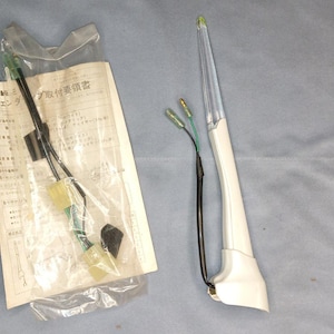 Op de afbeelding: Een witte en grijze autoantenne met een zwarte draad en een groene draad. De antenne is verpakt in een doorzichtige plastic zak met een witte instructiekaart. De instructiekaart is in het Japans en bevat de tekst "Instructies voor het installeren van de antenne".