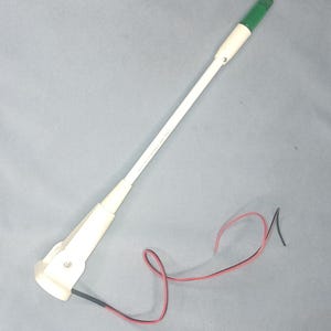May include: A white, elongated device with a green tip and a red and black wire. The device has a cylindrical body and a flared base. The wire is curled. The device is on a gray surface.