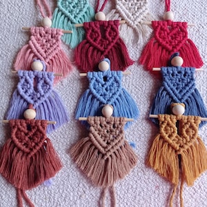 Può includere: Un set di dieci ornamenti per bambole in macramé con perline di legno e frange. Le bambole sono di vari colori, tra cui rosso, blu, rosa, marrone e bianco.