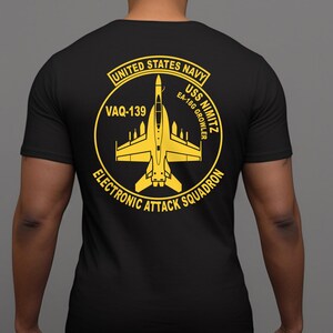 May include: Black t-shirt featuring a yellow graphic of a jet plane inside a circular design. The design includes the text "UNITED STATES NAVY", "VAQ-139", "USS NIMITZ", "EA-18G GROWLER", and "ELECTRONIC ATTACK SQUADRON".