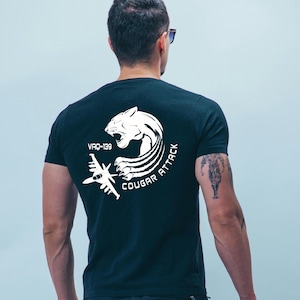 May include: Black t-shirt with a white graphic featuring a cougar, jet, and the text "COUGAR ATTACK" and "VRQ-139". The design is centered on the back of the shirt.