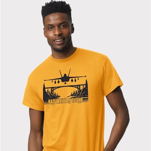 May include: Mustard yellow t-shirt featuring a black graphic of an airplane flying over a bridge, with the text "NAS Whidbey Island" below. The shirt is made of a soft material.