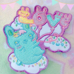 Pode incluir: Patch bordado com um coelho turquesa segurando balões que formam a palavra "LOVE". O patch tem uma base de nuvem branca com granulado colorido e uma borda roxa. A palavra "KAWAII GOODS" está na parte inferior.