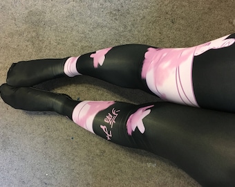 May include: Black leggings featuring a pink graphic print of a stylized character and Japanese text. The design includes a wide pink band across the thigh and lower leg areas, with a smooth, fitted fabric construction covering the feet.
