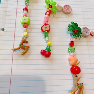 May include: Three handmade hair clips with decorative elements. Each clip features a string of beads and charms, including apples, cherries, deer, and ladybugs. The clips are adorned with buttons and various colorful accents.