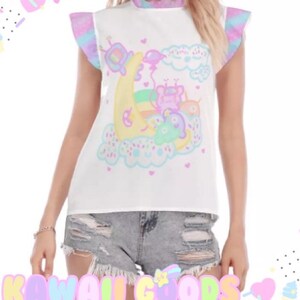 May include: White sleeveless top featuring a pastel cartoon graphic of a moon, clouds, and characters. The top has ruffled sleeves with pastel colours and a ruffled neckline. The text "KAWAII GOODS" is at the bottom.