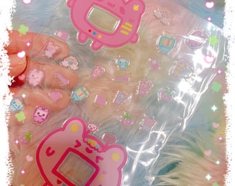 May include: A clear plastic sheet filled with stickers featuring a pink, cartoon-style handheld game console design. Additional stickers include various small, colorful graphics of food, stars, and other cute elements. The background is a soft, multi-colored fabric.
