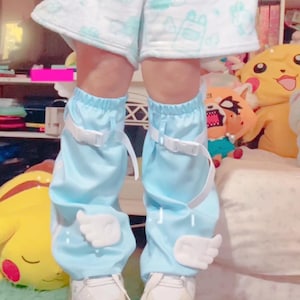 May include: Light blue leg warmers with white angel wing accents and white buckle straps. The leg warmers are worn over white platform sneakers with iridescent cloud designs. The shorts are white with light blue cloud patterns.
