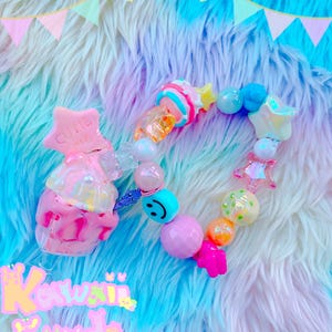 Könnte beinhalten: Ein farbenfrohes Perlenarmband mit einem rosa-weißen Eiscreme-Anhänger und einem Stern-Anhänger mit der Aufschrift "Cute Cake". Das Armband liegt auf einem flauschigen blau-rosa Hintergrund.