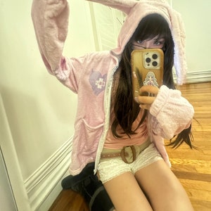 May include: A light pink hooded jacket with long bunny ears, a zipper, and pockets. The jacket has a heart-shaped graphic with a cross on the chest. The person is wearing a pink top, white shorts, and a brown belt.