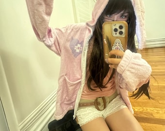 May include: A light pink hooded jacket with long bunny ears, a zipper, and pockets. The jacket has a heart-shaped graphic with a cross on the chest. The person is wearing a pink top, white shorts, and a brown belt.