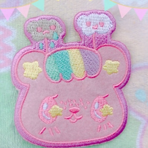 May include: A pink embroidered patch with a cute cartoon animal face. The animal has two clouds on its head, one with a sad face and one with a happy face. The clouds are raining rainbow colors.