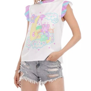 May include: White sleeveless top with a whimsical cartoon design featuring a crescent moon, clouds, and colourful creatures. The top has ruffled sleeves with a tie-dye pattern in shades of blue, purple, and pink. The design is in pastel colours.