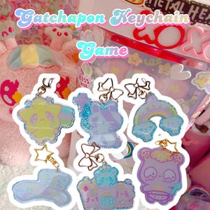 May include: Iridescent Gatchapon keyrings with cartoon characters and shapes, such as a bear, a bunny, and a rainbow. Each keyring has a gold-coloured bow or star charm. The text "Gatchapon Keychain Game" is visible.