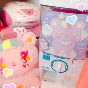May include: A selection of Kawaii-themed items, including a pink Hello Kitty CD player, a plush toy, and a gumball machine with a bear graphic. The gumball machine has a coin slot and a dispenser. The image has heart stickers and the text "KAWAII GOODS".