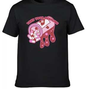 Peut inclure: T-shirt noir avec une illustration rose et blanche d'un flacon de m&eacute;dicaments et de pilules. Le texte "TAKE YOUR MEDICINE" est au-dessus du flacon. Le motif comprend des &eacute;toiles roses et un effet d&eacute;goulinant.