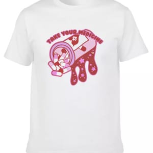 Peut inclure: T-shirt blanc avec un motif rose et rouge d'un flacon de m&eacute;dicaments d&eacute;versant des pilules. Le texte "TAKE YOUR MEDICINE" est arqu&eacute; au-dessus du flacon en rose. Le motif comprend des &eacute;toiles et un c&oelig;ur.