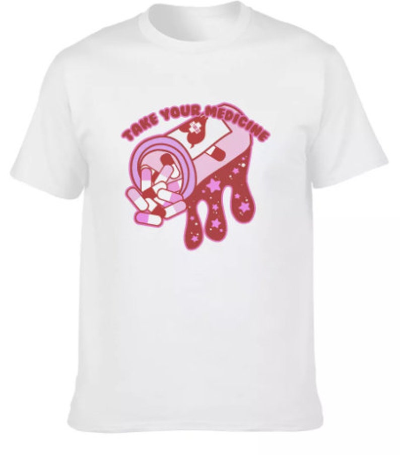 Peut inclure: T-shirt blanc avec un motif rose et rouge d'un flacon de m&eacute;dicaments d&eacute;versant des pilules. Le texte "TAKE YOUR MEDICINE" est arqu&eacute; au-dessus du flacon en rose. Le motif comprend des &eacute;toiles et un c&oelig;ur.