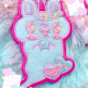 May include: A light blue bunny-shaped patch with a pink outline. The patch features embroidered details, including a crown, flowers, and the word "XOXO" on a heart. The background is a mix of pink, blue, and white.