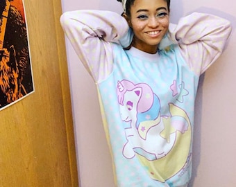 May include: A light blue sweatshirt with a unicorn graphic, stars, and a crescent moon. The sleeves are light pink with a white and light yellow star pattern. The person is wearing light blue tie-dye leggings and a white headband.