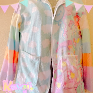 Könnte beinhalten: Eine pastellfarbene Kapuzen-Fleecejacke mit Reißverschluss. Die Jacke hat auf der einen Seite ein Wolkenmuster und auf der anderen Seite ein Cartoon-Charakter-Design. Sie hat zwei Vordertaschen und den Text "Kawaii Goods" unten.