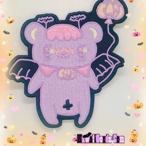 May include: Purple embroidered iron-on patch of a cute cartoon bear with wings, wearing a pink collar and holding a pumpkin balloon. The bear has a sparkly face and a cross on its chest.