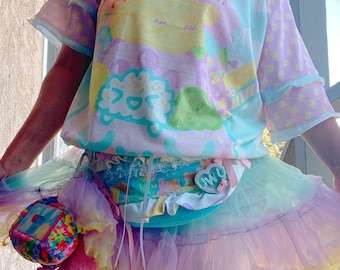 May include: A pastel-coloured outfit featuring a short-sleeved top with a whimsical design, a layered tulle skirt, and star-patterned leggings. The outfit includes a small, colourful bag and a belt with the text "OWU".