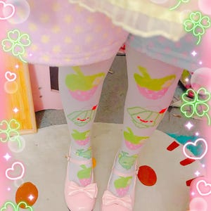 Op de afbeelding: Witte panty met een herhalend patroon van lichtgroene en roze aardbeivormige figuurtjes met kleine rode accenten. De panty wordt gecombineerd met lichtroze lakleren Mary Jane-schoenen met een strik aan de voorkant en een bandje.