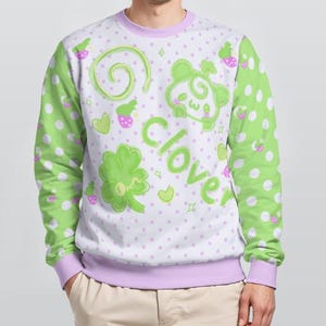 May include: A long-sleeve sweatshirt featuring a light purple base with white polka dots and green sleeves with white polka dots. The front displays a green four-leaf clover, a stylized green character, and the word clover in green text.