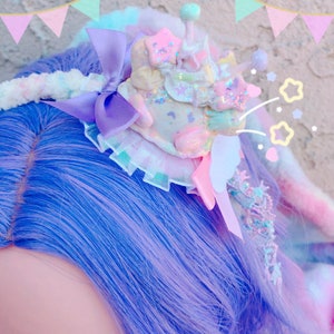 Puede incluir: Una peluca azul pastel y morada con una pinza para el pelo caprichosa que presenta un diseño de malvavisco blanco, rosa y azul con purpurina y estrellas. La pinza para el pelo está decorada con un lazo morado y un borde de encaje blanco.