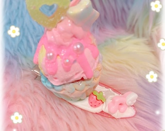 Puede incluir: Una pinza para el pelo de colores pastel que se asemeja a un cucurucho de helado caprichoso. La pinza presenta capas rosas, azules y amarillas, adornada con un caramelo en forma de corazón, perlas y una fresa. Sobre un fondo esponjoso y arcoíris.