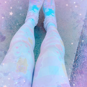 May include: A pair of white leggings with a pastel blue and pink pattern featuring cartoon bears. The leggings are worn with white socks and blue and white trainers. The background is a blurry image of a light blue and pink sky with white stars.