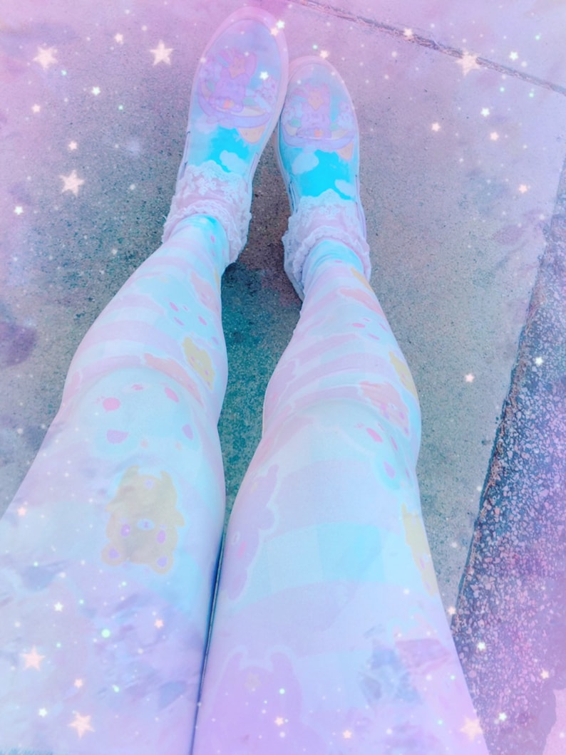 May include: A pair of white leggings with a pastel blue and pink pattern featuring cartoon bears. The leggings are worn with white socks and blue and white trainers. The background is a blurry image of a light blue and pink sky with white stars.