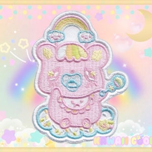 Peut inclure: Un patch brodé représentant un personnage de dessin animé rose portant un chapeau arc-en-ciel et tenant un hochet. Le personnage est entouré de nuages et d'étoiles. Le patch est sur un fond rose et bleu avec le texte "Kawaii Goods" en bas.