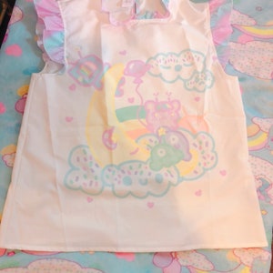 May include: A white sleeveless top featuring a pastel illustration of a bear on a crescent moon, with clouds and balloons. The top has frilled sleeves and a frilled collar. The text "KAWAII GOODS" is at the bottom.