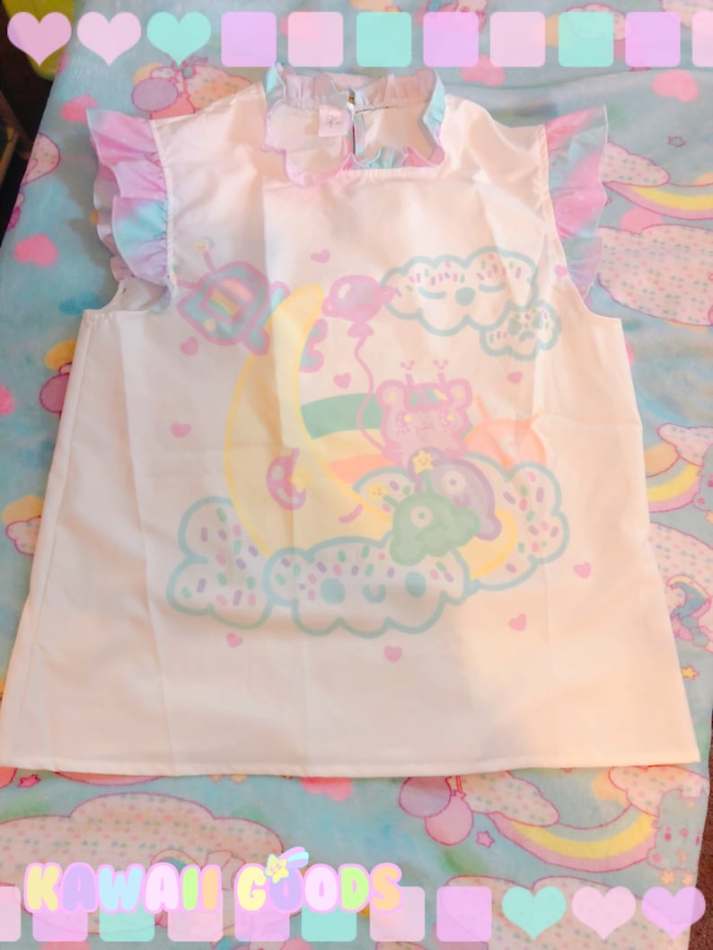 May include: A white sleeveless top featuring a pastel illustration of a bear on a crescent moon, with clouds and balloons. The top has frilled sleeves and a frilled collar. The text "KAWAII GOODS" is at the bottom.