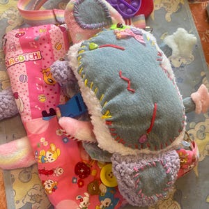 May include: A plush toy with a denim patch on its back, featuring colorful hand-stitched details, attached to a pink fabric bag with a repeating cartoon print and the text MAGOTCHI. A purple silicone sensory toy is visible near the top strap.