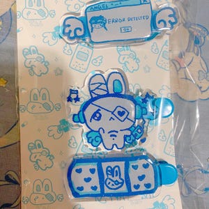 May include: A pack of three clear acrylic hair clips with blue and white illustrations. The top clip reads "ANGEL A-A" and "ERROR DETECTED". The middle clip features a bunny character with headphones. The bottom clip has a bunny and heart design.
