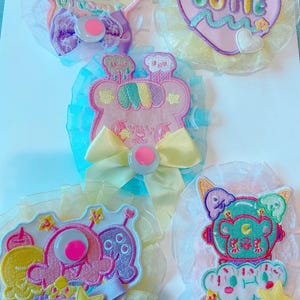 May include: Five colourful embroidered patches with text and cartoon designs. The patches feature a star, heart, and alien, with text that reads "Sweet Dreams" and "Cutie". Each patch has a button and ribbon accents.