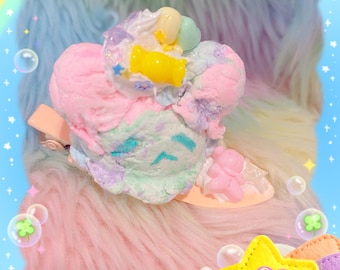 Puede incluir: Una pinza para el pelo de colores pastel que se asemeja a una bola de helado, adornada con chispas y caramelos. La pinza presenta tonos rosas, azules y verdes, con un pequeño acento en forma de oso. El fondo es una textura esponjosa de colores arcoíris.