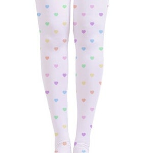 Op de afbeelding: Witte dijhoge sokken met een patroon van kleine, pastelkleurige hartjes in geel, groen, paars en roze. De sokken worden van de voeten naar boven getoond en reiken tot halverwege de dij.