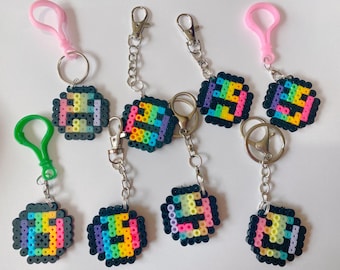 Puede incluir: Una colección de llaveros de pixel art con un diseño de arcoíris. Cada llavero presenta una cara sonriente y colorida hecha de pequeñas cuentas redondas. Los llaveros tienen anillos y cierres de metal plateado, algunos con clips de plástico rosa o verde.