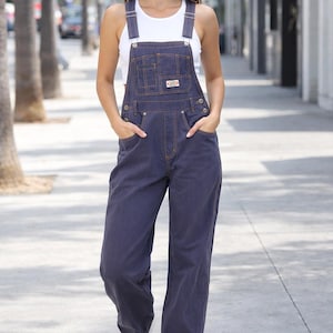 May include: A person wearing blue denim overalls with a white tank top and white sneakers. The overalls have visible stitching and pockets. The person is walking on a sidewalk.