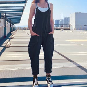 May include: Black jumpsuit with a relaxed fit, featuring a scoop neckline and pockets. The model is wearing a white tank top underneath and black and white sneakers. The jumpsuit has a casual, comfortable style, suitable for everyday wear.