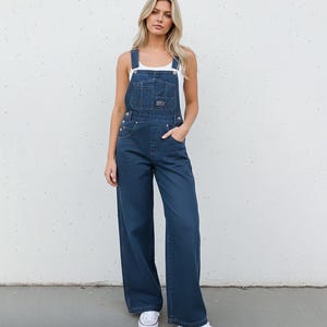 May include: Dark blue denim overalls with wide legs. The overalls feature a bib pocket, adjustable straps, and side button closures. The model is wearing a white tank top and white sneakers. The overall design is casual and comfortable.