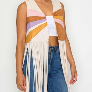 May include: A sleeveless vest with a colorful sunburst design. The vest features a cream-colored base with pink, purple, orange, and brown suede-like panels. Long cream-colored fringe hangs from the bottom of the vest. The vest is open in the front.