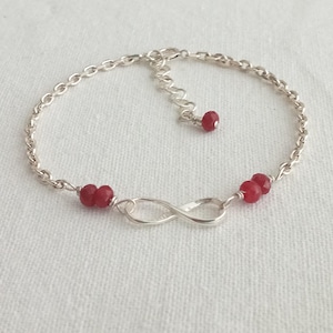 Op de afbeelding: Een zilveren armband met een oneindigheidsbedel en drie rode edelsteen kralen.