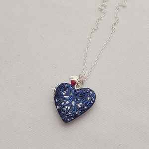 Puede incluir: Un collar de cadena plateada con un relicario en forma de corazón y una capa de filigrana azul oscuro. El relicario está decorado con una pequeña cuenta roja y un diminuto dije de mano de Hamsa plateado unido a la argolla.