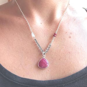 May include: A silver necklace featuring a teardrop-shaped, faceted, pink gemstone pendant. The necklace has a delicate chain with small red and clear beads. The pendant is the focal point, showcasing the vibrant color and cut of the stone.
