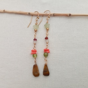 Puede incluir: Un par de aretes largos de oro con una variedad de cuentas de colores, incluyendo verde, rojo y naranja. Los aretes tienen una piedra de ojo de tigre en forma de lágrima en la parte inferior.